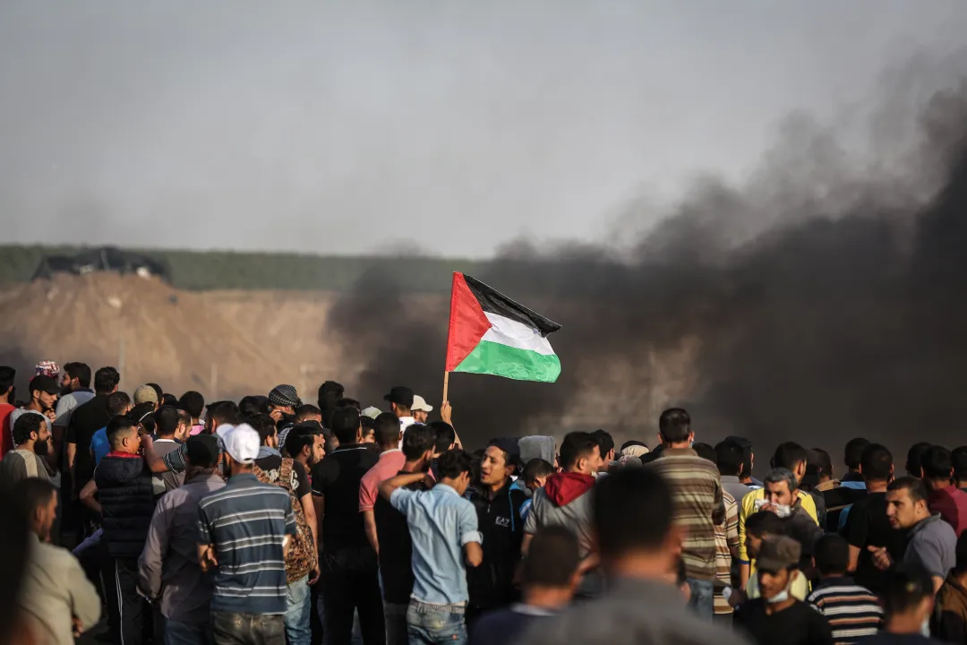 Kerumunan warga Palestina mengibarkan bendera nasional di tengah kepulan asap.