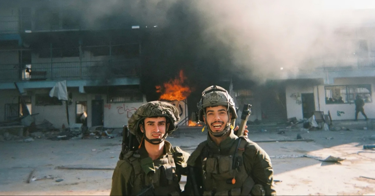 Dua tentara Israel berseragam berpose tersenyum di depan bangunan sekolah yang terbakar di Gaza.